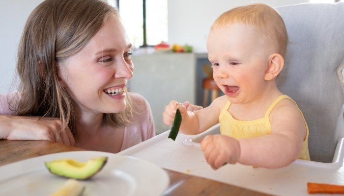 Joyful mom watching baby eating solid food in high chair.