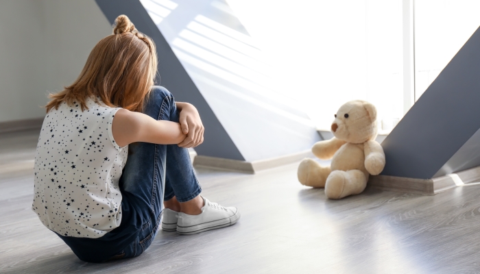 Little girl with teddy bear sitting on floor near window in empty room.