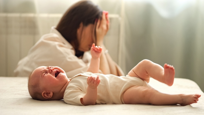 Tired infant girl cries on double bed against depressed mother sitting on floor.