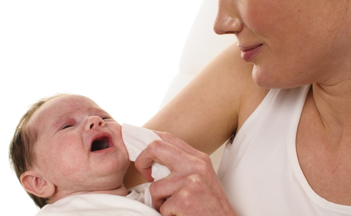 An adult woman with white shirt holding a also dressed in white infant tenderly in her arms and dabbed it with a tissue for cleaning the mouth.