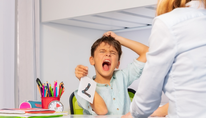 Boy with autism spectrum disorder tear hairs and scream expressing negative emotions while learning numbers sitting by teacher.