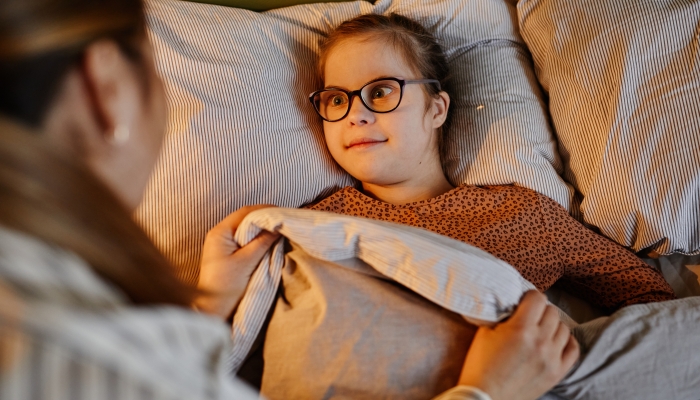 High angle portrait of caring mother tucking in child with down syndrome at bedtime in cozy lamp light, copy space.