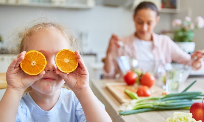 Playful blond girl covering eyes with orange slices in kitchen at home.