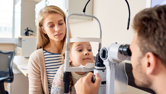 Portrait of cute little girl siting on mothers lap looking at slit lamp machine during medical check up in eye clinic.