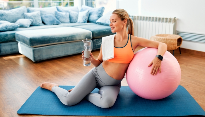 A young pregnant woman in sportswear sitting with a fitness ball on a rubber mat and drinking water after a workout.