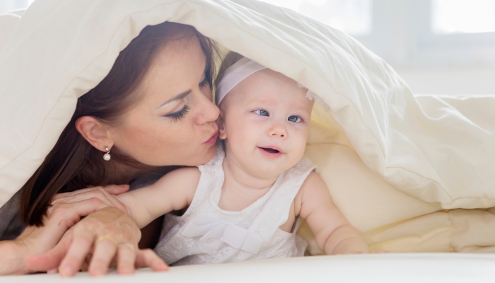 Close up of young mother kissing her baby girl while lying under a blanket in the bedroom.