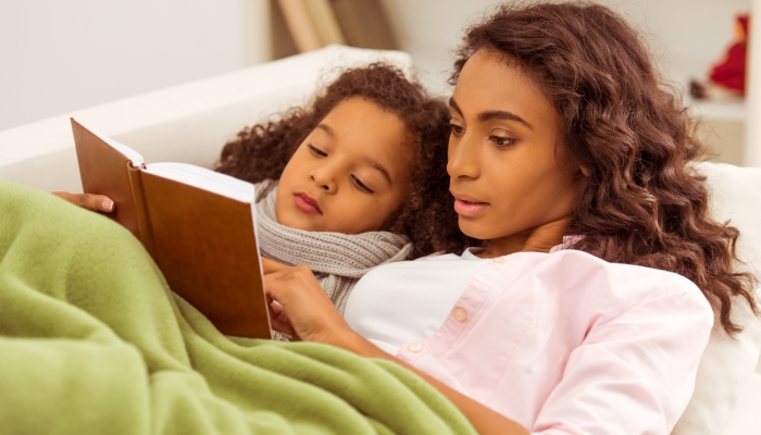 Cute little Afro-American girl with scarf on her neck is ill, lying in bed with her mother.