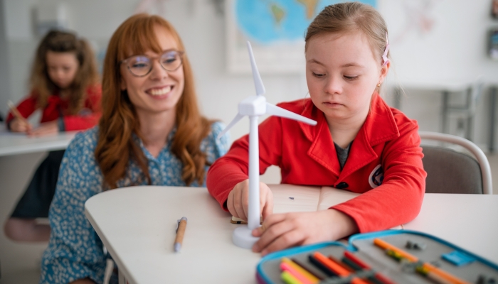 Down syndrome schoolgirl with model of wind turbine with help of teacher learning about eco-friendly renewable sources of energy in class at school.
