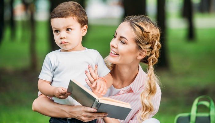 Happy mother with sad little son reading book at park.