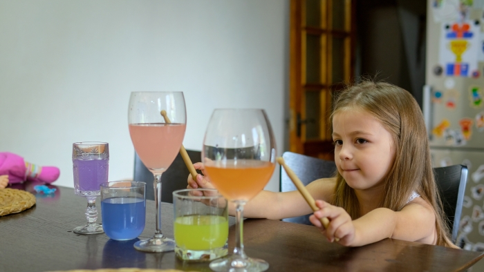 Little girl, toddler plays music with drumsticks in glasses of water in the kitchen.
