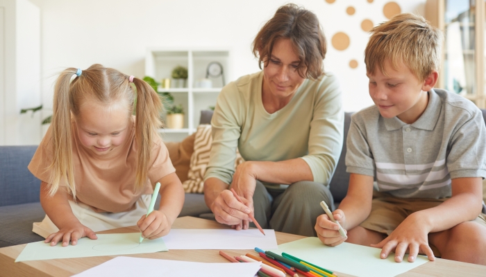 Portrait of cute blonde girl with down syndrome drawing with mother and brother together in home interior.