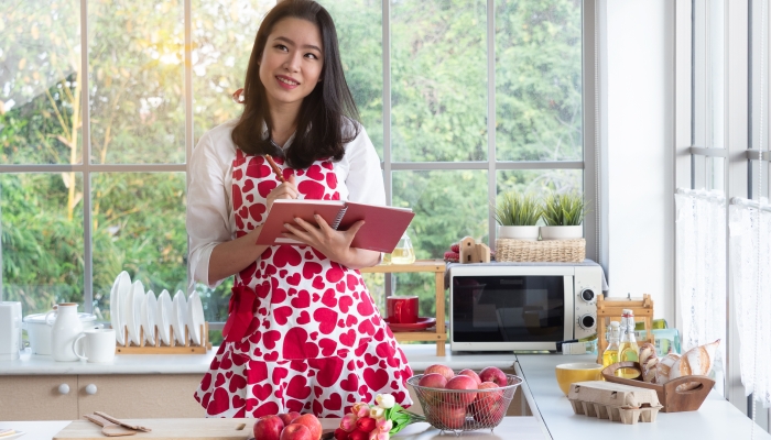 Pretty Asian woman wearing cute apron holding cook book thinking of what to cook.