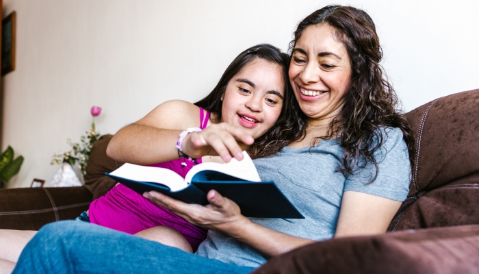 Smiling latin teen girl with down syndrome and her mom reading a book at home.