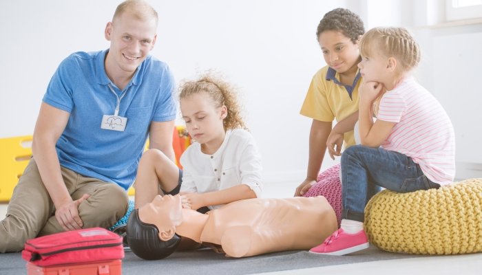 Smiling rescuer and children learning first aid on a phantom in a room with red kit.