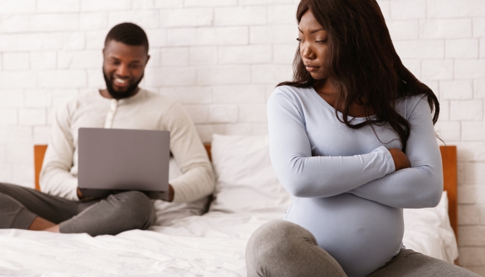 Stressed pregnant afro woman sitting on bed while her husband using laptop, family conflicts during pregnancy.