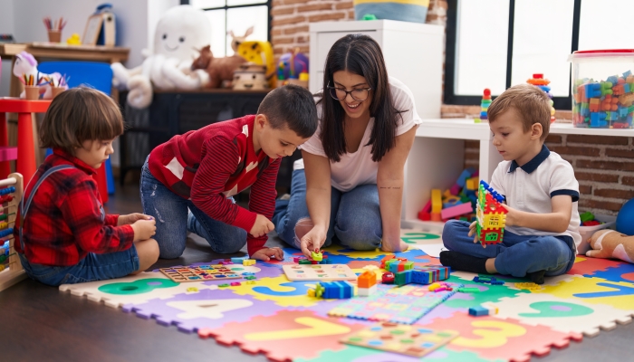 Teacher with group of boys playing with maths puzzle game sitting on floor at kindergarten.