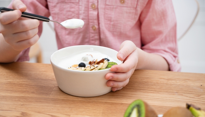 The hands of a girl in a pink shirt hold a bowl of yogurt, granola and fresh fruit on a wooden table.