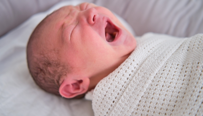 A newborn baby is crying swaddled on the bed.