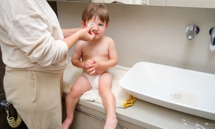 In the bathroom at home, a woman is taking care of her child's eye hygiene by cleaning them with a cotton pad.