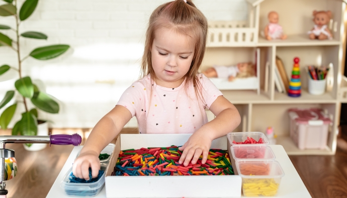 Little girl toddler playing with sensory bin with colored dyed pasta.