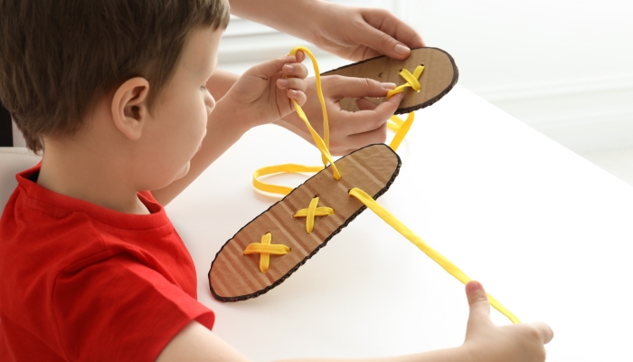 Mother teaching son to tie shoe laces using training cardboard template at white table.