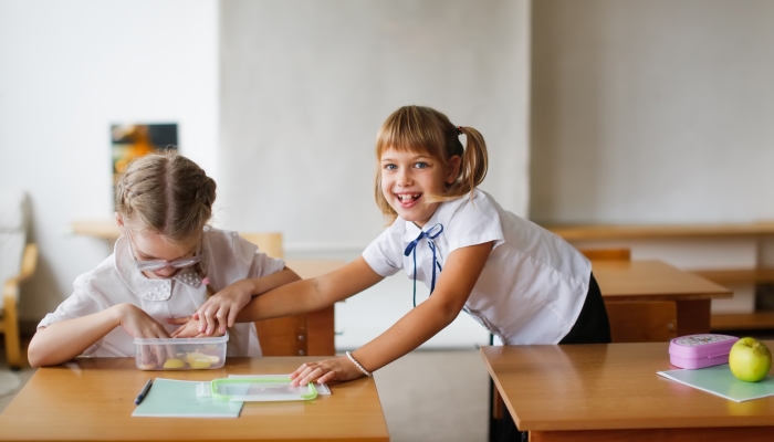 Two girlfriends of classmate at school have snack at desk.