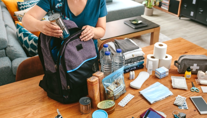 Unrecognizable woman putting cans of food to prepare emergency backpack in living room.