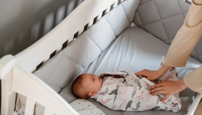 White young woman in housecoat swaddling her newborn baby in crib at bedroom.