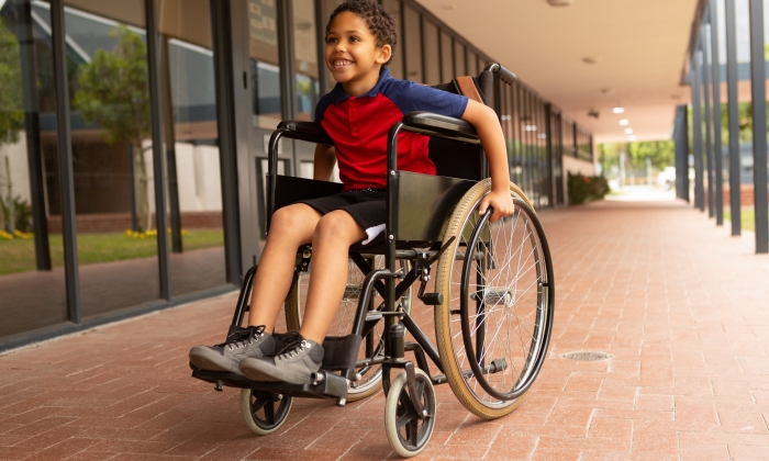 Front view of happy mixed-race disabled schoolboy sitting on wheelchair in corridor at elementary school.