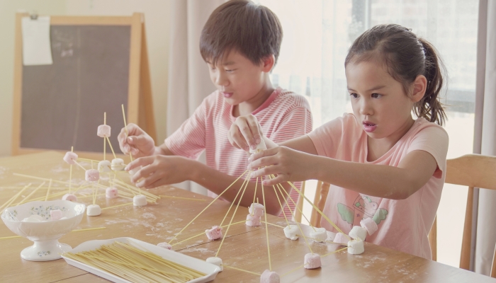 Mixed race young Asian children building tower with spaghetti and marshmallow learning remotely at home.