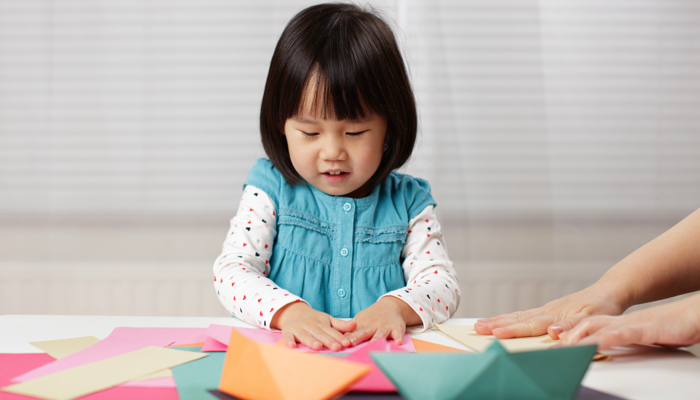 A little girl sitting at a table folding origami paper.