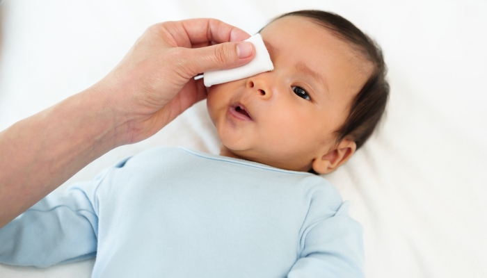 Mother cleaning and wiping newborn baby eye with cotton pad on a bed.