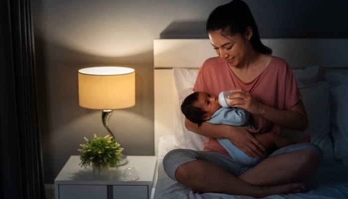 Mother feeding milk bottle to her newborn baby on a bed at night.