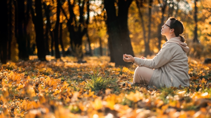 Caucasian middle aged woman meditating in lotus pose at autumn park with sunlight.