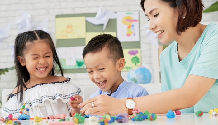 Cheerful Asian teacher playing with boy and girl with plasticine figurines in art class.