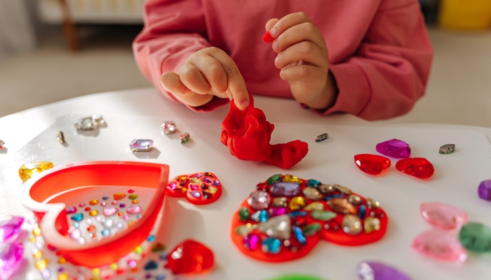 Child hands creating red heart from play dough for modeling with decorate from crystal rhinestones and shiny stones.