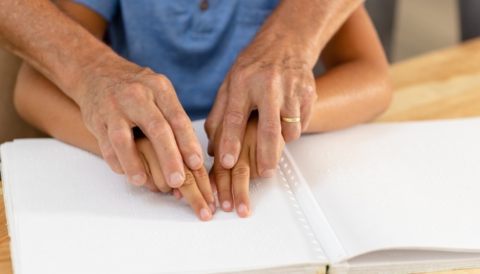 Cropped hands of caucasian grandfather assisting blind grandson in reading braille book at home.