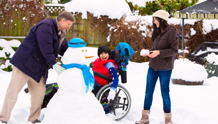 Disabled biracial young boy in wheelchair building a snowman with father and sister during winter after heavy snowfall.