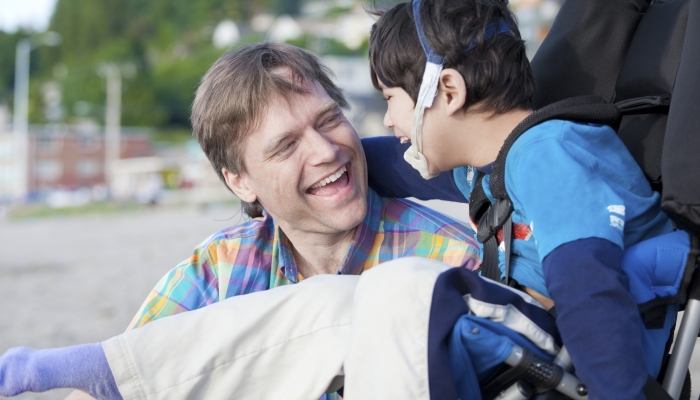 Father and disabled five year old son laughing together on beach.