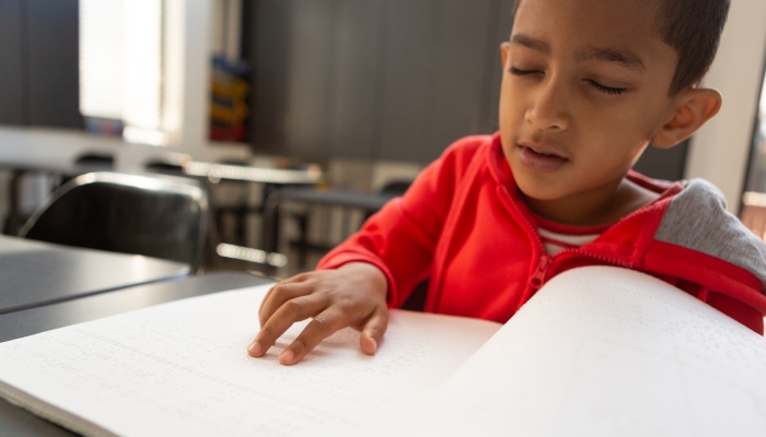 Front view of blind mixed-race schoolboy reading a braille book at desk in a classroom at elementary school.