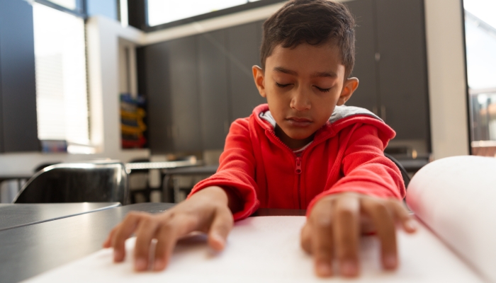 Front view of cute blind mixed-race schoolboy reading a braille book at desk in a classroom.