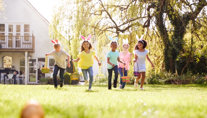 Group Of Children Wearing Bunny Ears Running To Pick Up Chocolate Egg On Easter Egg Hunt In Garden.