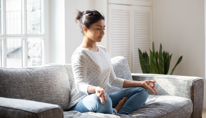 Indian female sitting on couch lotus pose put hands on lap folded fingers closed eyes enjoy meditation.