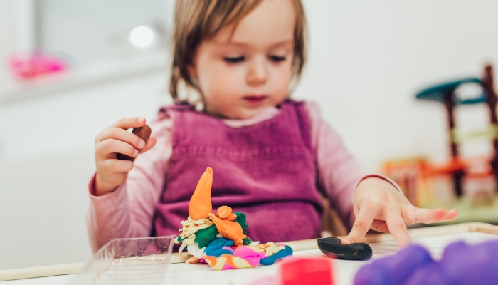 Kid girl is playing with plasticine while sitting at table in nursery room.