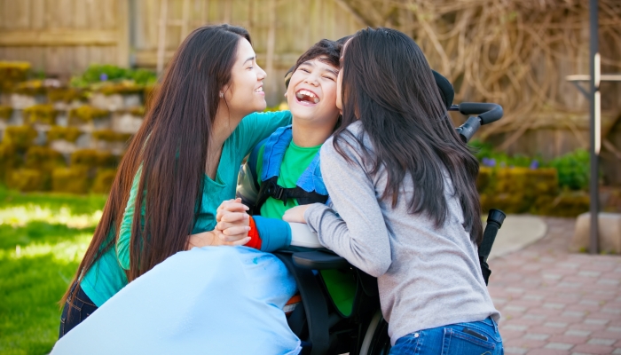Laughing disabled boy in wheelchair being hugged and kissed by two teenage girls on patio outdoors.
