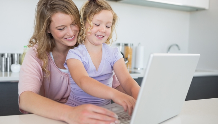 Mother assisting daughter in using laptop at table in kitchen.