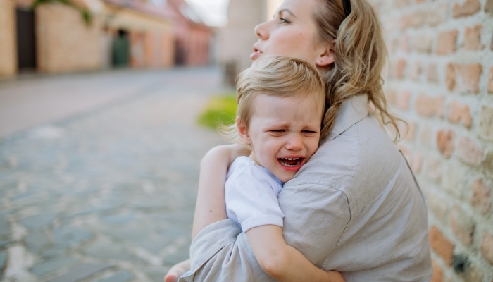 Mother consling her little daughter crying, holding her in arms in street in summer.