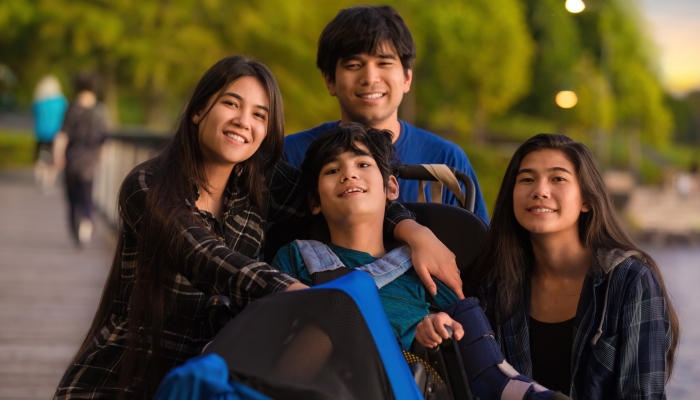 Older siblings, brother and sisters, surrounding little disabled boy in wheelchair outdoors at lake park.