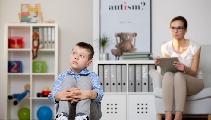 Psychologist in glasses is looking at kid sick of autism sitting on carpet in classroom.