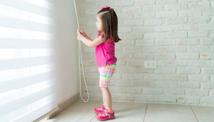 Side view of small girl opening blinds of window at home.